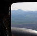 A military personnel member is seated inside an aircraft, viewed in silhouette, looking out through a large open window or door at a mountainous landscape below. The scene appears to be taken during an aerial mission over a remote, rugged terrain with rolling hills and valleys. A cylindrical metallic object, likely part of the aircraft's exterior or a fuel tank, is visible in the lower portion of the frame.