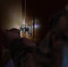 A close-up photograph showing a cat's paw reaching toward or touching a cabinet door hinge. The image is taken from inside or very close to a wooden cabinet, with a blurred cat visible in the foreground. The focus is centered on a standard European-style concealed cabinet hinge mounted on a wooden door.