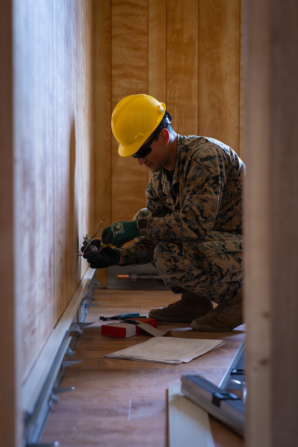 A military service member in digital camouflage uniform is performing electrical work, crouching near a wall outlet or electrical box inside a wooden-paneled structure. The individual is wearing a yellow hard hat, safety glasses, and green work gloves while using hand tools. Reference documents and various tools are laid out on the floor nearby.
