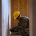 A military service member in digital camouflage uniform is performing electrical work, crouching near a wall outlet or electrical box inside a wooden-paneled structure. The individual is wearing a yellow hard hat, safety glasses, and green work gloves while using hand tools. Reference documents and various tools are laid out on the floor nearby.