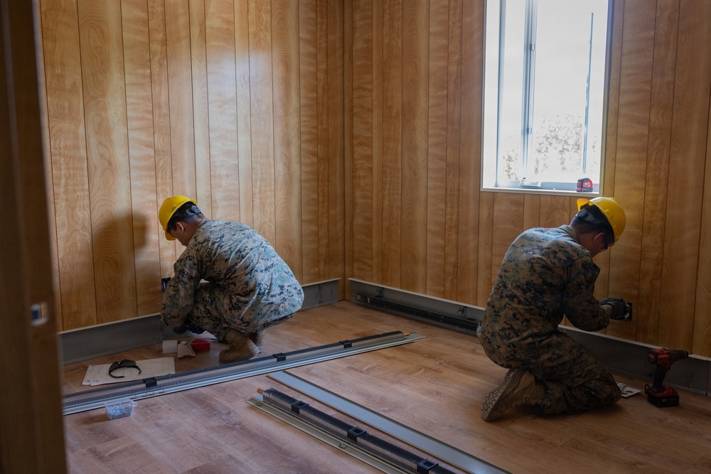 Two military personnel in camouflage uniforms and yellow hard hats are performing interior construction or renovation work inside a room with wood-paneled walls and laminate flooring. They appear to be installing floor trim or baseboard components near the walls. The setting suggests a military construction or engineering project.