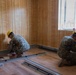 Two military personnel in camouflage uniforms and yellow hard hats are performing interior construction or renovation work inside a room with wood-paneled walls and laminate flooring. They appear to be installing floor trim or baseboard components near the walls. The setting suggests a military construction or engineering project.