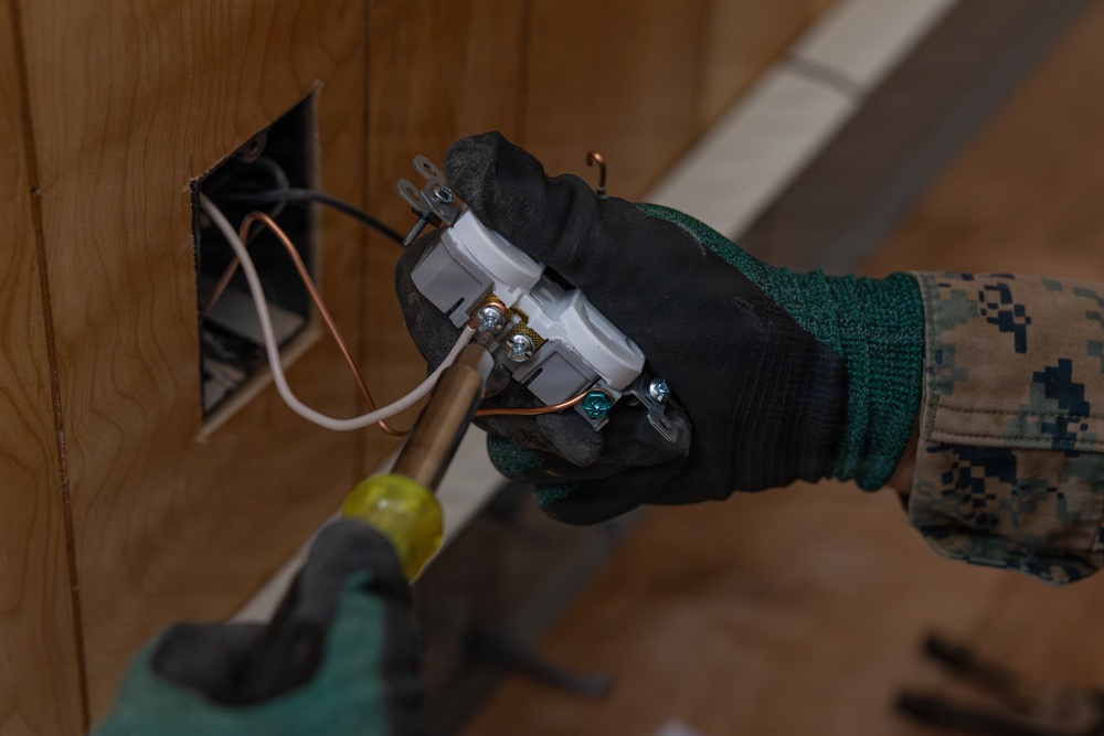 A person wearing military camouflage uniform and black/green work gloves is installing or removing an electrical outlet from a wall box. The worker is using a screwdriver to work on the outlet, which has visible copper wiring including hot, neutral, and ground wires. The wall appears to be plywood or wood paneling with a standard electrical junction box.