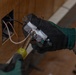 A person wearing military camouflage uniform and black/green work gloves is installing or removing an electrical outlet from a wall box. The worker is using a screwdriver to work on the outlet, which has visible copper wiring including hot, neutral, and ground wires. The wall appears to be plywood or wood paneling with a standard electrical junction box.