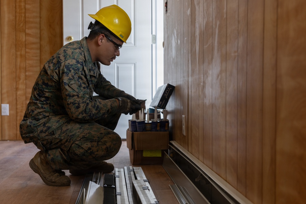 A U.S. Marine in digital camouflage uniform and yellow hard hat is crouched down performing construction or repair work along the base of a wood-paneled interior wall. The service member is working with supplies from an open cardboard box containing what appear to be caulk or adhesive tubes. The setting appears to be the interior of a building undergoing renovation or repair work.