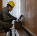 A U.S. Marine in digital camouflage uniform and yellow hard hat is crouched down performing construction or repair work along the base of a wood-paneled interior wall. The service member is working with supplies from an open cardboard box containing what appear to be caulk or adhesive tubes. The setting appears to be the interior of a building undergoing renovation or repair work.