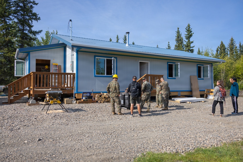 A group of military personnel in camouflage uniforms, along with two civilians, are gathered outside a small single-story building with a blue metal roof and white siding. The building appears to be under construction or renovation, with building materials, a wood pile, and a miter saw visible on the gravel lot. The setting appears to be a rural or remote area surrounded by conifer and birch trees, consistent with an Alaskan environment.