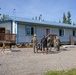 A group of military personnel in camouflage uniforms, along with two civilians, are gathered outside a small single-story building with a blue metal roof and white siding. The building appears to be under construction or renovation, with building materials, a wood pile, and a miter saw visible on the gravel lot. The setting appears to be a rural or remote area surrounded by conifer and birch trees, consistent with an Alaskan environment.