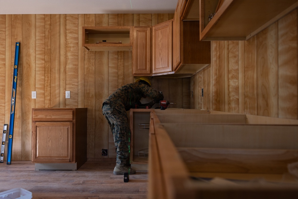 A person wearing military camouflage uniform and a yellow hard hat is installing kitchen cabinets in a room with wood-paneled walls. The space appears to be under renovation, with upper and lower cabinets partially installed and tools visible nearby. The setting suggests a construction or home improvement project, possibly on a military base.