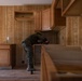 A person wearing military camouflage uniform and a yellow hard hat is installing kitchen cabinets in a room with wood-paneled walls. The space appears to be under renovation, with upper and lower cabinets partially installed and tools visible nearby. The setting suggests a construction or home improvement project, possibly on a military base.