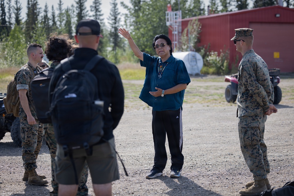 A civilian individual in a blue shirt and black Adidas track pants appears to be briefing or directing a group of U.S. Marines in military camouflage uniforms outdoors. The setting appears to be a gravel area near a red building surrounded by coniferous trees, suggesting a northern or Alaskan environment. The civilian is gesturing with one raised hand, indicating active communication or instruction.