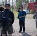 A civilian individual in a blue shirt and black Adidas track pants appears to be briefing or directing a group of U.S. Marines in military camouflage uniforms outdoors. The setting appears to be a gravel area near a red building surrounded by coniferous trees, suggesting a northern or Alaskan environment. The civilian is gesturing with one raised hand, indicating active communication or instruction.