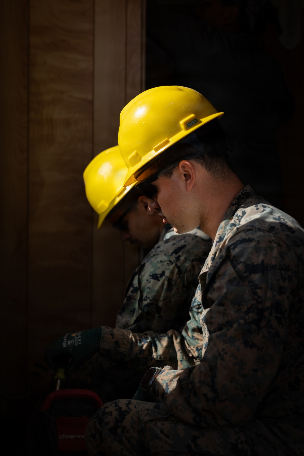 Two individuals wearing US Marine Corps MARPAT camouflage uniforms and yellow MSA hard hats are seated together in a dimly lit area, likely engaged in a construction or engineering task. Both wear work gloves, and one appears to be holding a tool. The dramatic side lighting creates strong contrast, highlighting their profiles against a dark background.