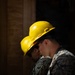 Two individuals wearing US Marine Corps MARPAT camouflage uniforms and yellow MSA hard hats are seated together in a dimly lit area, likely engaged in a construction or engineering task. Both wear work gloves, and one appears to be holding a tool. The dramatic side lighting creates strong contrast, highlighting their profiles against a dark background.