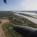 An aerial photograph taken from a helicopter showing a small rural settlement situated between a dirt/gravel airstrip on the left and a wide, silt-laden river on the right. The community consists of scattered buildings surrounded by boreal forest, consistent with a remote subarctic or Alaskan village. Helicopter components including a rotor blade and engine nacelle are visible in the foreground, confirming the aerial vantage point.