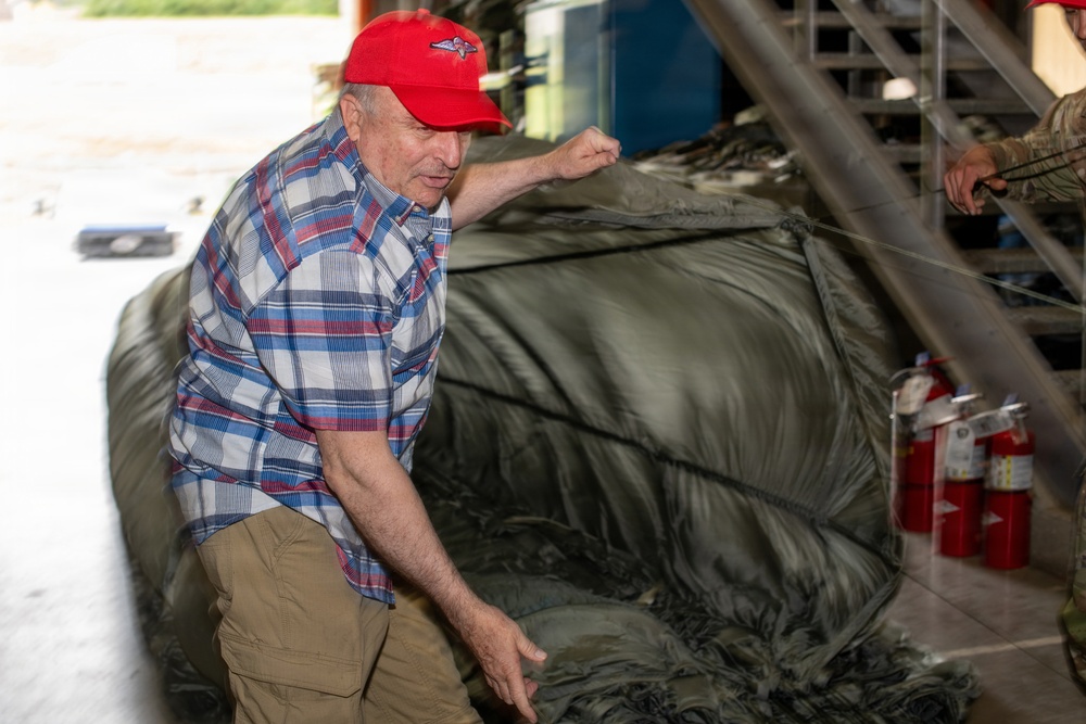 An older man wearing a red baseball cap and plaid shirt is examining or handling a large olive-green military-style inflatable boat or raft inside what appears to be a hangar or storage facility. He is leaning over the deflated or partially inflated craft, inspecting it closely. A second person in camouflage clothing is partially visible on the right side of the frame.