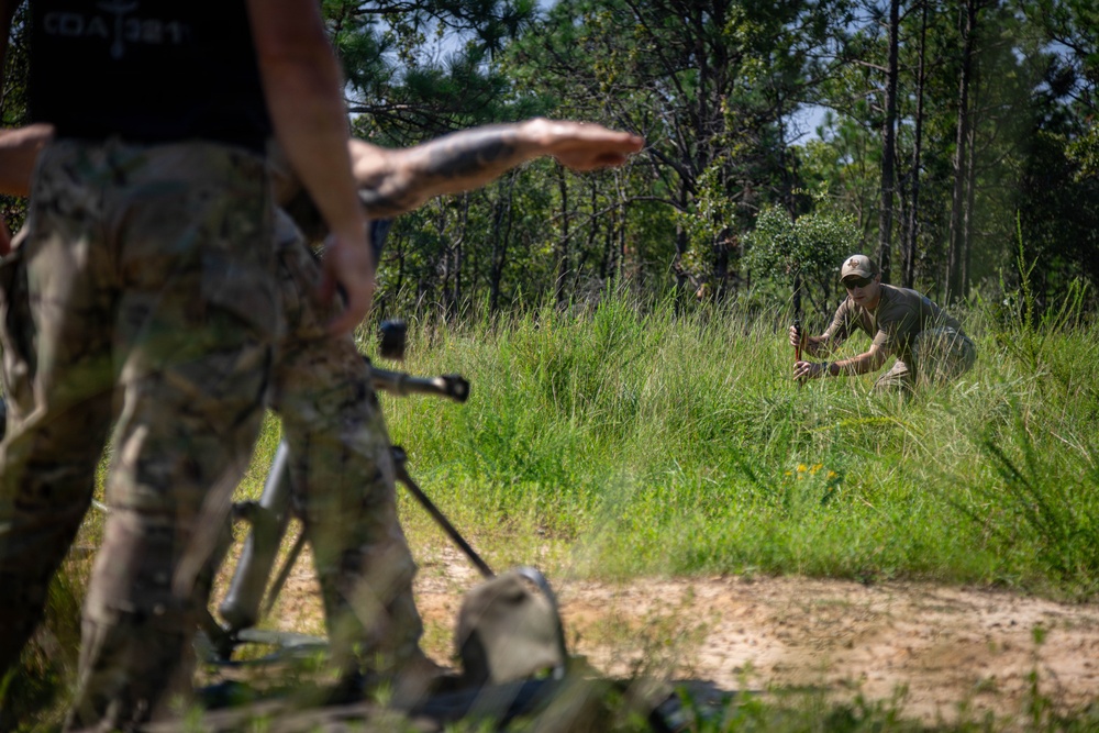 The image shows what appears to be a military or tactical training exercise in a wooded outdoor environment. A person in the foreground wearing camouflage pants holds a weapon and gestures with their hand, while a second individual in the background crouches in tall grass. The setting suggests a field training scenario with sandy terrain and dense tree line.