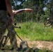 The image shows what appears to be a military or tactical training exercise in a wooded outdoor environment. A person in the foreground wearing camouflage pants holds a weapon and gestures with their hand, while a second individual in the background crouches in tall grass. The setting suggests a field training scenario with sandy terrain and dense tree line.