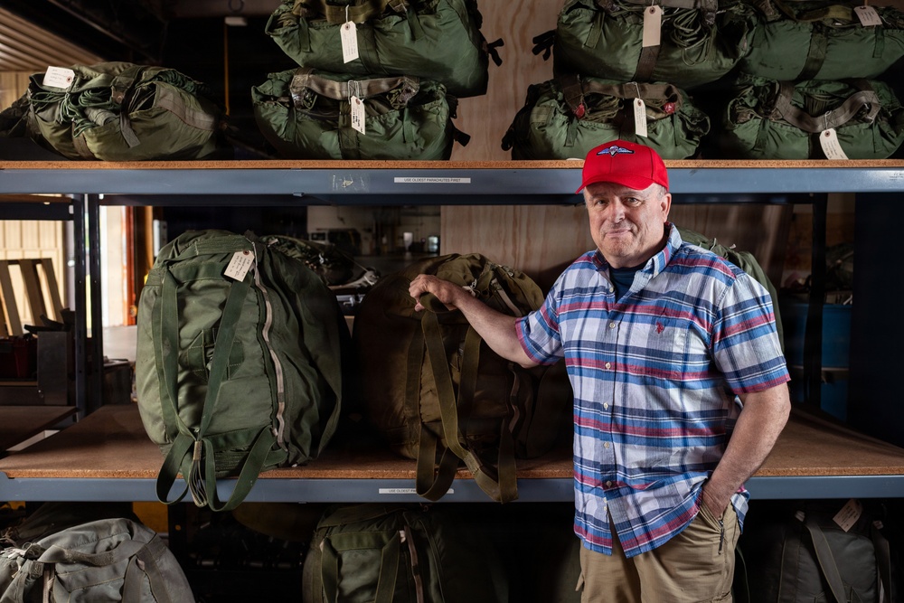 A middle-aged man wearing a red baseball cap with a parachutist/airborne insignia and a plaid shirt stands in front of industrial shelving units stocked with olive drab military-style duffel bags and backpacks. The bags appear to be tagged with identification labels, suggesting an organized storage or collection facility. The setting resembles a military surplus warehouse or a parachute/equipment storage room.