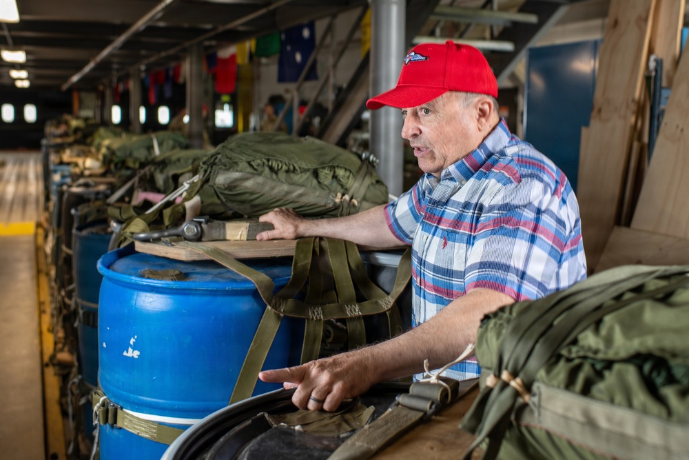 An older man wearing a red baseball cap with a wing/parachute emblem and a plaid shirt is gesturing and speaking while standing among military equipment. He appears to be giving a tour or explanation of the displayed gear, which includes olive drab military rucksacks, blue barrels, and olive webbing straps. The setting appears to be the interior of a military vessel or museum exhibit, with international signal flags visible in the background.