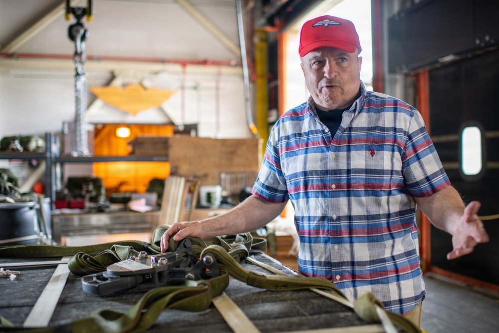 A middle-aged man wearing a red cap with a wing/parachute emblem and a plaid shirt stands in what appears to be a hangar or workshop. He is gesturing with his right hand while his left hand rests on military-style olive drab straps and harness equipment laid out on a table. The background contains various equipment, shelving, and what appears to be an orange parachute or canopy hanging from the ceiling.