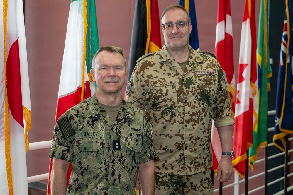 Two military personnel in uniform are posing together in front of a display of international flags. The individual on the left wears a U.S. Navy digital camouflage uniform with the name tape 'BAILEY', while the individual on the right wears a German Bundeswehr flecktarn camouflage uniform with the name tape 'STOCKMANN'. The setting appears to be an official military or NATO facility.