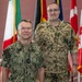 Two military personnel in uniform are posing together in front of a display of international flags. The individual on the left wears a U.S. Navy digital camouflage uniform with the name tape 'BAILEY', while the individual on the right wears a German Bundeswehr flecktarn camouflage uniform with the name tape 'STOCKMANN'. The setting appears to be an official military or NATO facility.