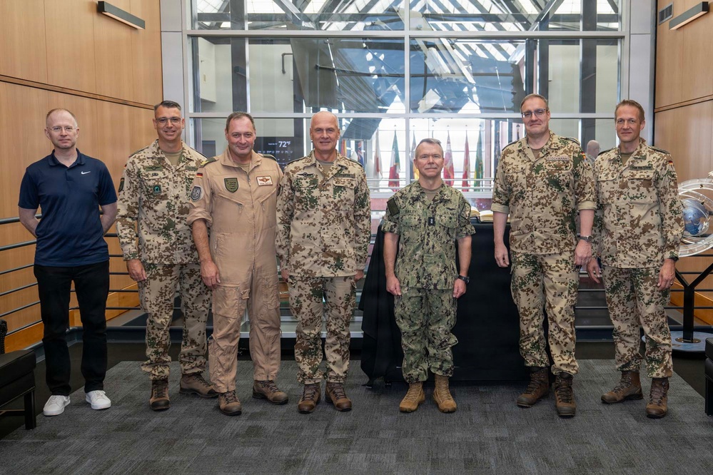 A group of seven individuals pose for a formal group photo in what appears to be a military headquarters or command facility. Six of the individuals are wearing military uniforms from what appear to be German Bundeswehr (flecktarn camouflage pattern) and one US Navy uniform, while one civilian in a navy blue Nike polo stands on the far left. Multiple national flags are visible in the background, suggesting a multinational military setting.
