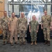 A group of seven individuals pose for a formal group photo in what appears to be a military headquarters or command facility. Six of the individuals are wearing military uniforms from what appear to be German Bundeswehr (flecktarn camouflage pattern) and one US Navy uniform, while one civilian in a navy blue Nike polo stands on the far left. Multiple national flags are visible in the background, suggesting a multinational military setting.