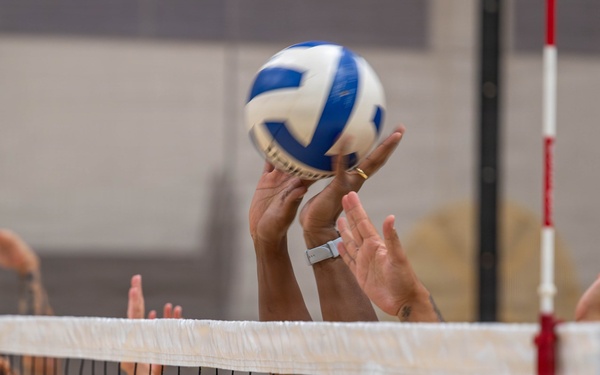 A close-up action shot of a volleyball match showing players at the net during a blocking or setting play. Multiple players in blue uniforms are reaching above the white net toward a blue and white volleyball. The image captures an intense moment of competition with hands and arms prominently featured near the ball.
