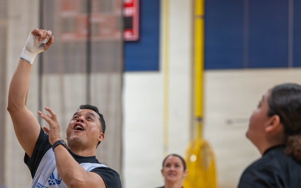 A group of wheelchair basketball players are competing or practicing in an indoor gymnasium. The central figure, wearing a 'Wounded Warrior' vest, is shooting or celebrating with his arm raised. Other players wearing 'ARMY' shirts are visible in the background, suggesting this is a Wounded Warrior Project adaptive sports event.