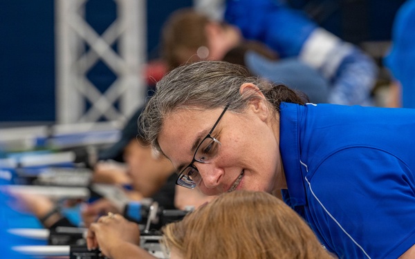 An indoor shooting range session where an adult instructor in a blue polo shirt and glasses is leaning over to coach or assist a young participant with red/auburn hair. Multiple participants are lined up along a shooting bench, each positioned behind what appear to be air rifles or precision rifles. The setting appears to be an organized youth or competitive shooting program based on the uniform blue attire and professional equipment.