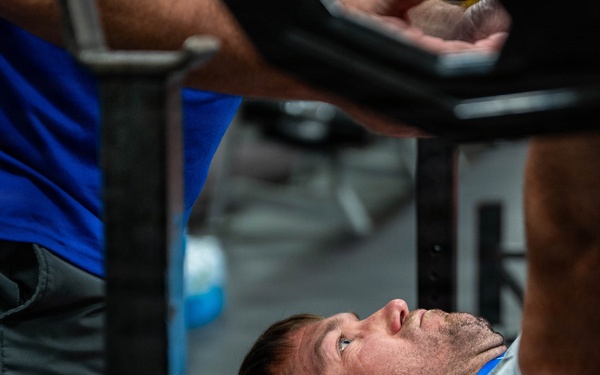 A man is lying on a red weight bench performing or preparing to perform a bench press exercise in a gym setting. A spotter wearing a blue shirt stands nearby, and a loaded barbell is visible in the upper portion of the frame. The close-up angle emphasizes the lifter's focused expression and the intensity of the exercise.