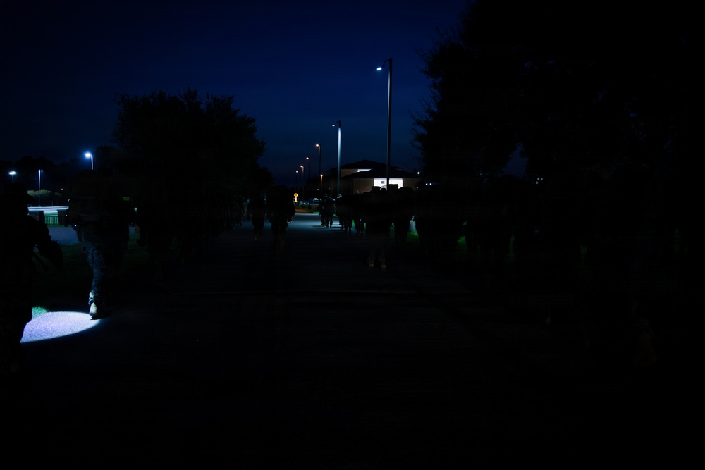 A nighttime outdoor scene showing a group of people walking along a path or road in low-light conditions. Several street lights illuminate the area, with trees silhouetted against a dark blue sky. A small pool of light is visible on the lower left, possibly from a handheld flashlight or ground-level light source.
