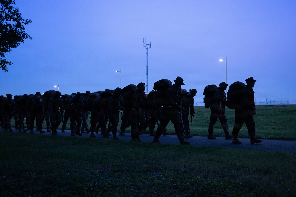 A large column of military personnel conducting a ruck march along a paved path during early morning or dusk conditions. The soldiers are silhouetted against a blue twilight sky, carrying large rucksacks and wearing combat gear. The formation stretches back along the path with street lights and a communications tower visible in the background.