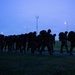 A large column of military personnel conducting a ruck march along a paved path during early morning or dusk conditions. The soldiers are silhouetted against a blue twilight sky, carrying large rucksacks and wearing combat gear. The formation stretches back along the path with street lights and a communications tower visible in the background.