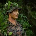 A U.S. Marine identified by the name tape 'GARDNER' is photographed in a jungle or forested environment wearing full camouflage gear. He has applied face paint in green and brown tones and has tucked natural foliage into his boonie hat for additional concealment. The image appears to document field training or a tactical exercise focused on camouflage and concealment techniques.