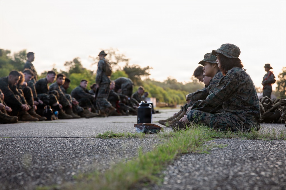 A group of military personnel in camouflage uniforms are seated on the ground along a paved surface, appearing to be in a rest or briefing period during a training exercise. The scene is captured at low angle during early morning or late afternoon light, with trees visible in the background. Two individuals in the foreground are prominently featured wearing MARPAT (Marine pattern) camouflage uniforms and boonie hats.