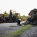 A group of military personnel in camouflage uniforms are seated on the ground along a paved surface, appearing to be in a rest or briefing period during a training exercise. The scene is captured at low angle during early morning or late afternoon light, with trees visible in the background. Two individuals in the foreground are prominently featured wearing MARPAT (Marine pattern) camouflage uniforms and boonie hats.