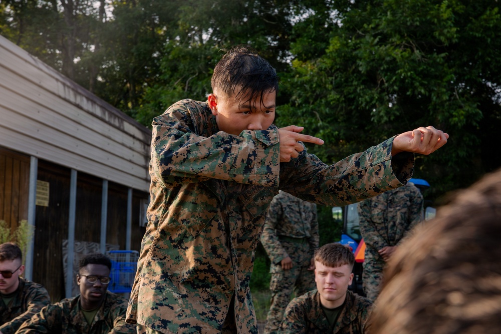 A group of military personnel in MARPAT (Marine pattern) camouflage uniforms are gathered outdoors near a wooden structure surrounded by trees. One individual in the foreground is standing and demonstrating a technique with outstretched arms, appearing to instruct or demonstrate to the seated group. The setting suggests a training exercise or field instruction session.