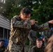 A group of military personnel in MARPAT (Marine pattern) camouflage uniforms are gathered outdoors near a wooden structure surrounded by trees. One individual in the foreground is standing and demonstrating a technique with outstretched arms, appearing to instruct or demonstrate to the seated group. The setting suggests a training exercise or field instruction session.