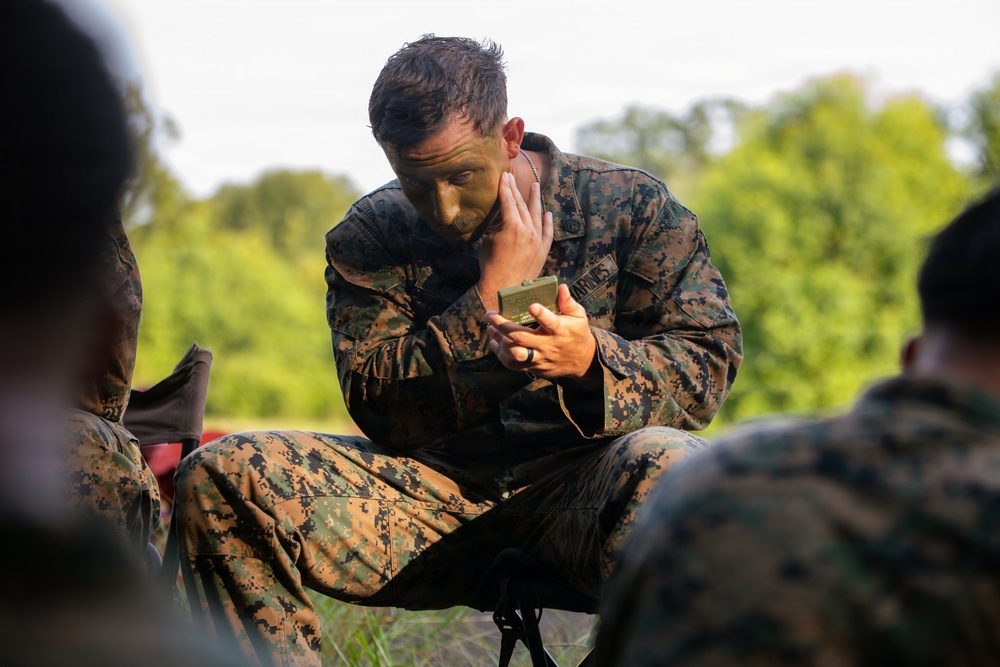 A U.S. Marine in MARPAT digital camouflage uniform is applying camouflage face paint using a small compact mirror while seated outdoors. The marine appears to be preparing for a field exercise or tactical operation. Other marines in similar uniforms are partially visible in the foreground and background.