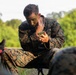 A U.S. Marine in MARPAT digital camouflage uniform is applying camouflage face paint using a small compact mirror while seated outdoors. The marine appears to be preparing for a field exercise or tactical operation. Other marines in similar uniforms are partially visible in the foreground and background.