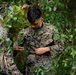 U.S. Marines in MARPAT camouflage uniforms are engaged in what appears to be a field training exercise involving vegetation and natural camouflage techniques. The central Marine, identified by a name tape reading 'MARTIN' and 'U.S. MARINES' on his uniform, is examining or attaching foliage to his gear. The setting is a dense, green wooded environment consistent with survival or field craft training.
