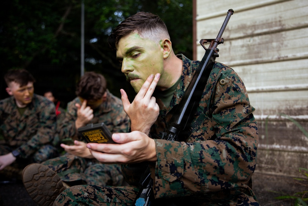 A U.S. Marine in MARPAT digital camouflage uniform is applying green camouflage face paint using a compact mirror while holding a rifle slung over his shoulder. Several other Marines in the background are similarly applying face paint in preparation for a field exercise. The setting appears to be an outdoor staging area near a wooden structure.