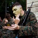 A U.S. Marine in MARPAT digital camouflage uniform is applying green camouflage face paint using a compact mirror while holding a rifle slung over his shoulder. Several other Marines in the background are similarly applying face paint in preparation for a field exercise. The setting appears to be an outdoor staging area near a wooden structure.