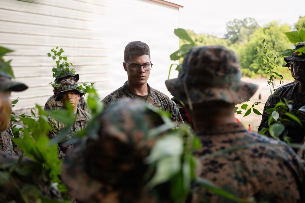 A group of military personnel in woodland camouflage uniforms are gathered in a vegetated outdoor area near a white building. A central figure wearing glasses and no hat appears to be speaking or briefing the others. The setting suggests a field training exercise or outdoor instruction session.