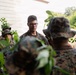 A group of military personnel in woodland camouflage uniforms are gathered in a vegetated outdoor area near a white building. A central figure wearing glasses and no hat appears to be speaking or briefing the others. The setting suggests a field training exercise or outdoor instruction session.