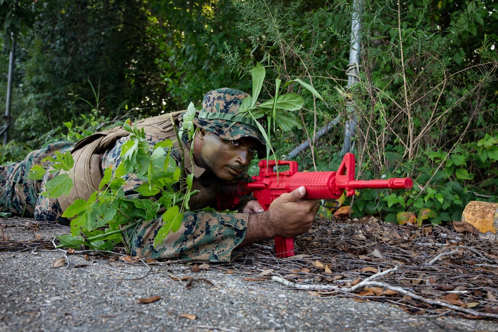 A military service member in MARPAT (Marine pattern) camouflage uniform is lying prone in a wooded area during what appears to be a training exercise. The individual is aiming a bright red training weapon, which is a non-functional replica used specifically for military training purposes to distinguish it from live firearms. The soldier has face paint applied and vegetation tucked into their gear for additional concealment.