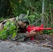 A military service member in MARPAT (Marine pattern) camouflage uniform is lying prone in a wooded area during what appears to be a training exercise. The individual is aiming a bright red training weapon, which is a non-functional replica used specifically for military training purposes to distinguish it from live firearms. The soldier has face paint applied and vegetation tucked into their gear for additional concealment.